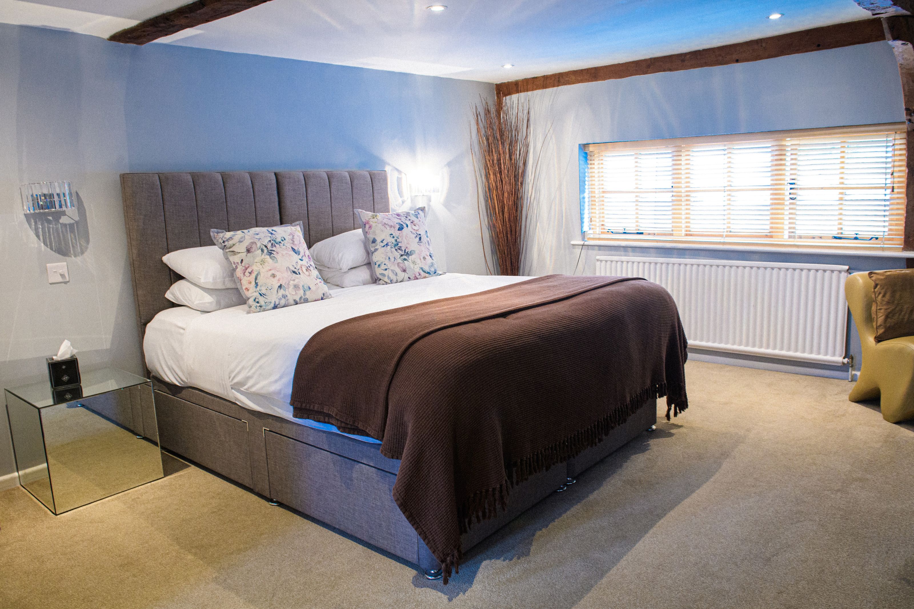 A boutique bedroom with a grey upholstered bed, floral cushions and a brown throw at Sheene Mill, Melbourn, Cambridgeshire
