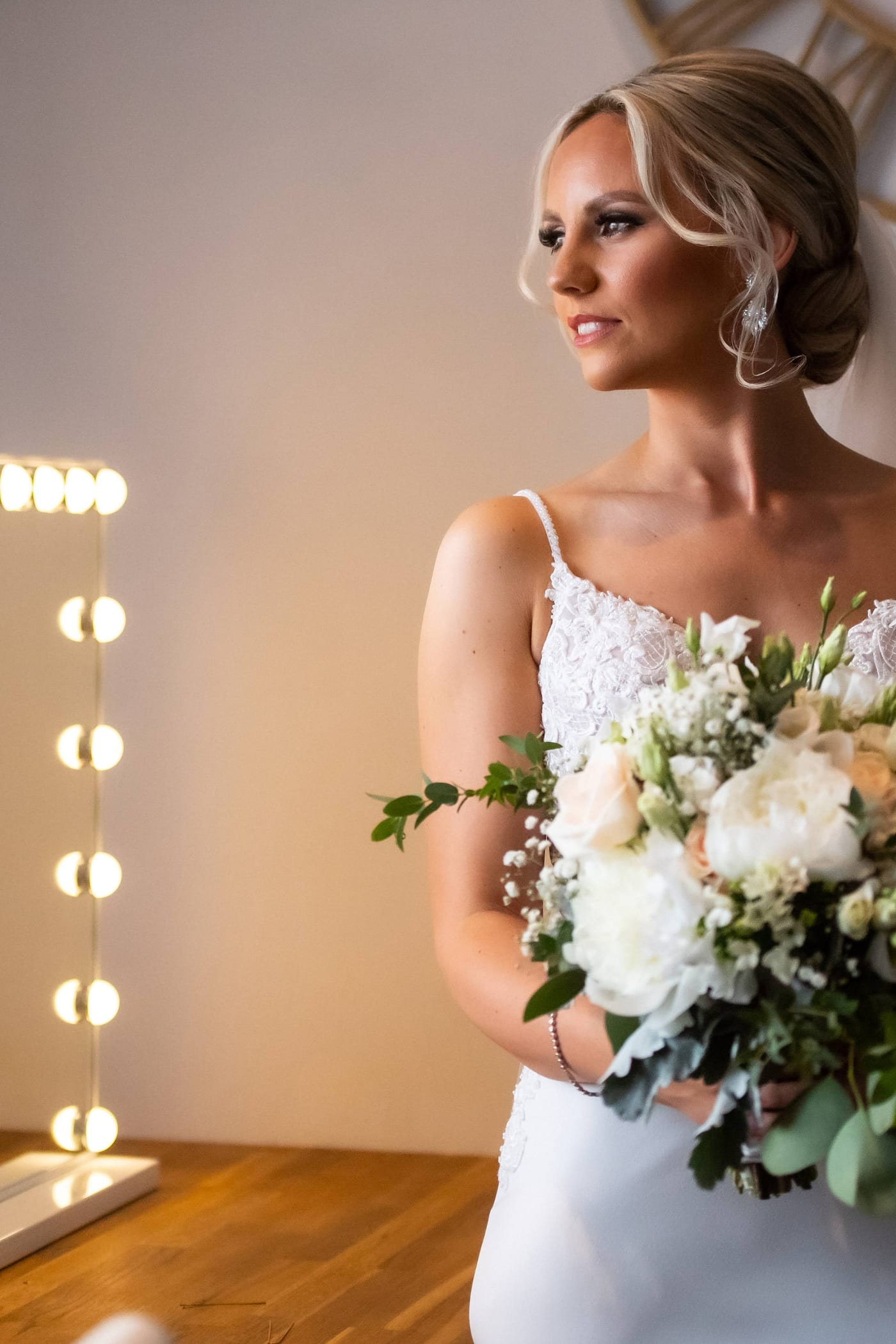 A bride holding her bouquet beside a lit vanity mirror before the ceremony at Sheene Mill