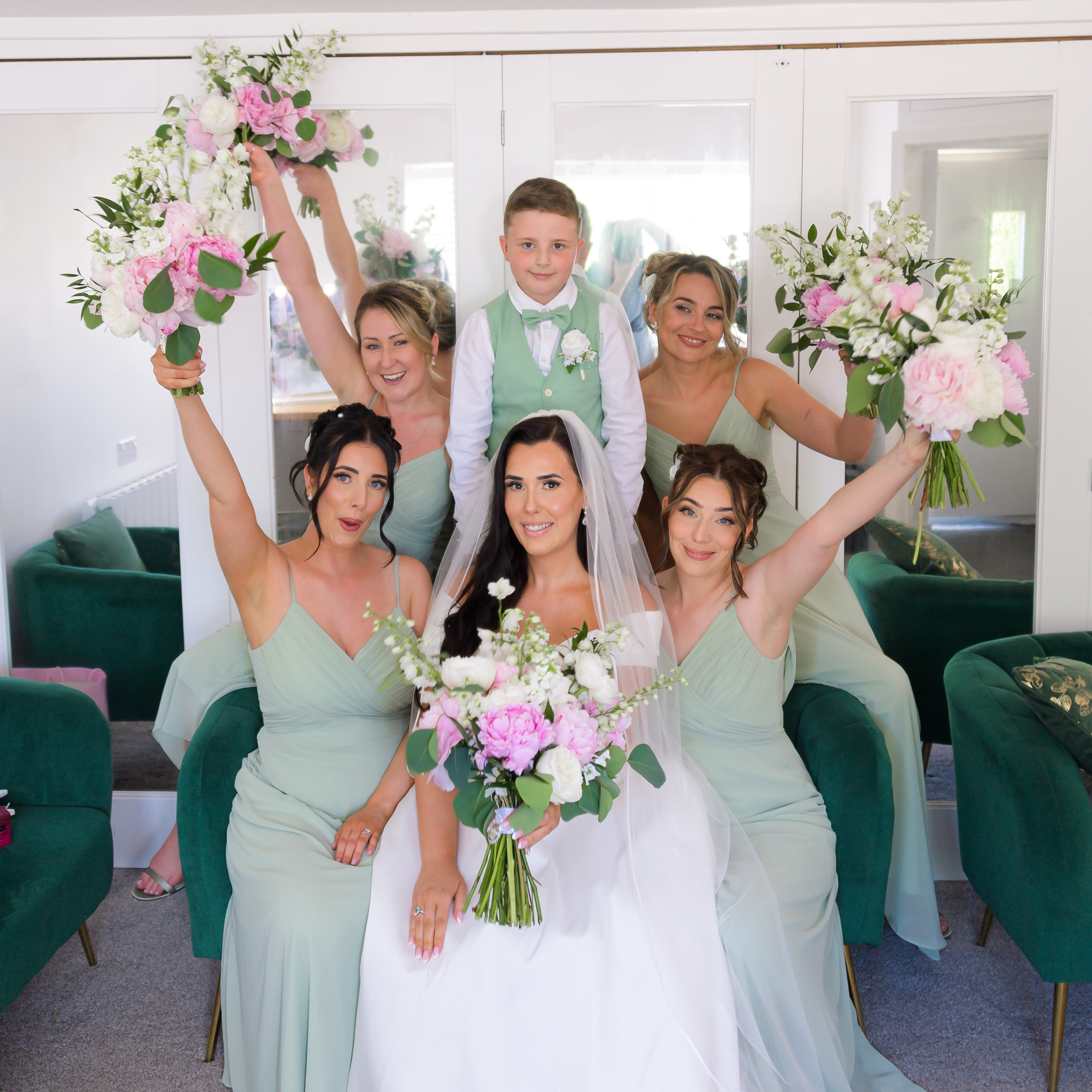 A bride seated with her bridesmaids in sage green, bouquets raised, before the ceremony at Sheene Mill
