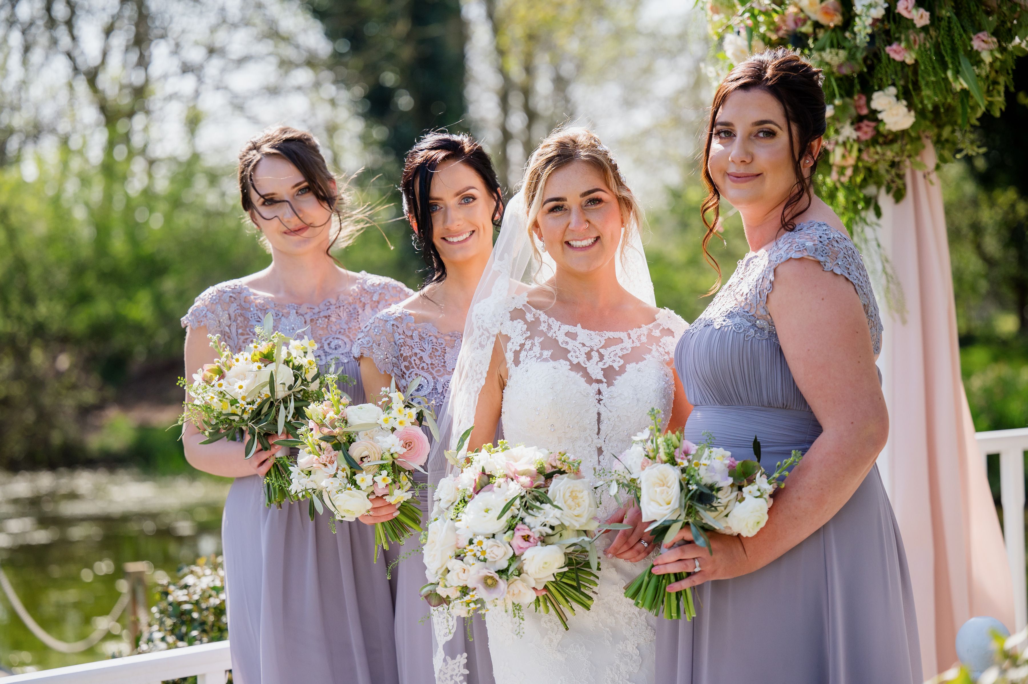 A bride and her three bridesmaids in lilac holding bouquets beside the mill pond at Sheene Mill, Cambridgeshire