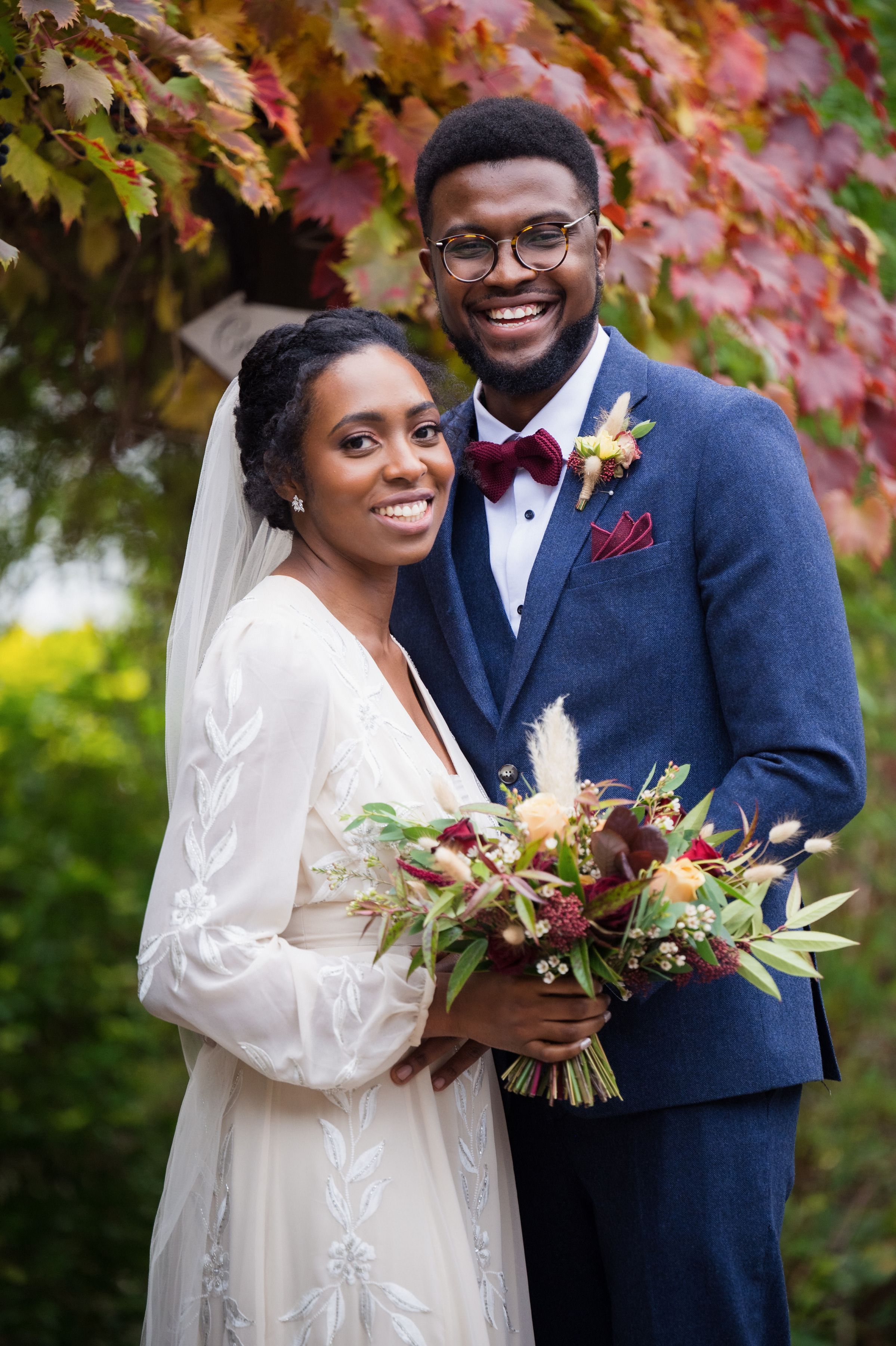 A bride and groom smiling with her bouquet in front of autumn leaves at Sheene Mill