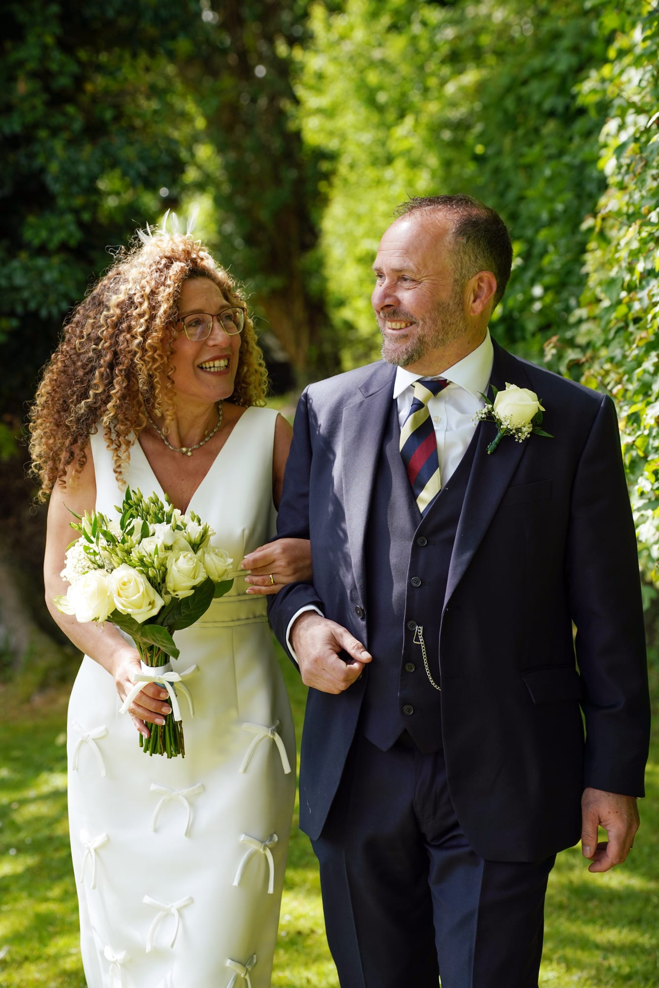 A bride and groom walking arm in arm through the sunlit gardens at Sheene Mill