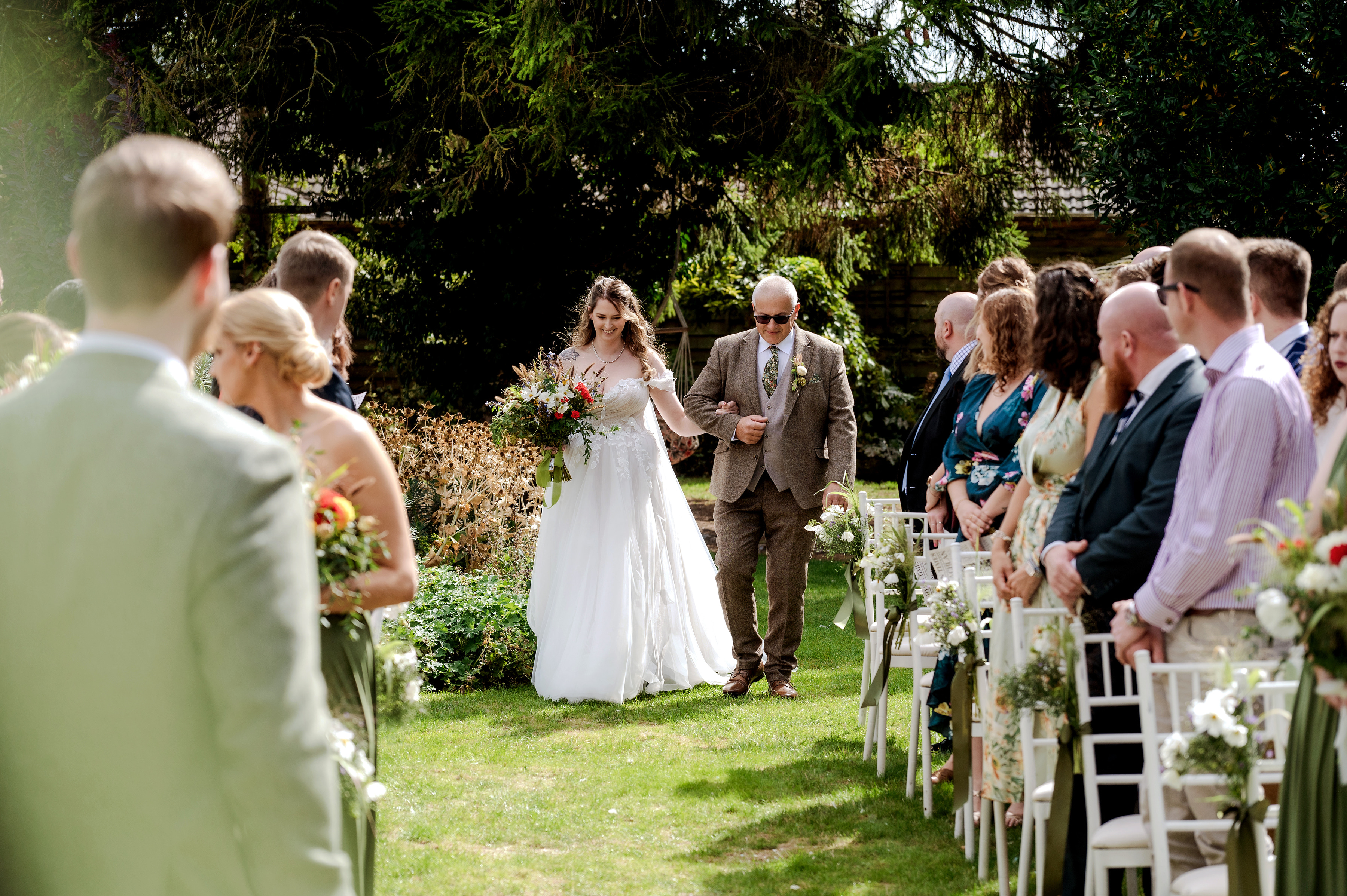 A bride walking down the outdoor aisle with her father as guests look on at Sheene Mill