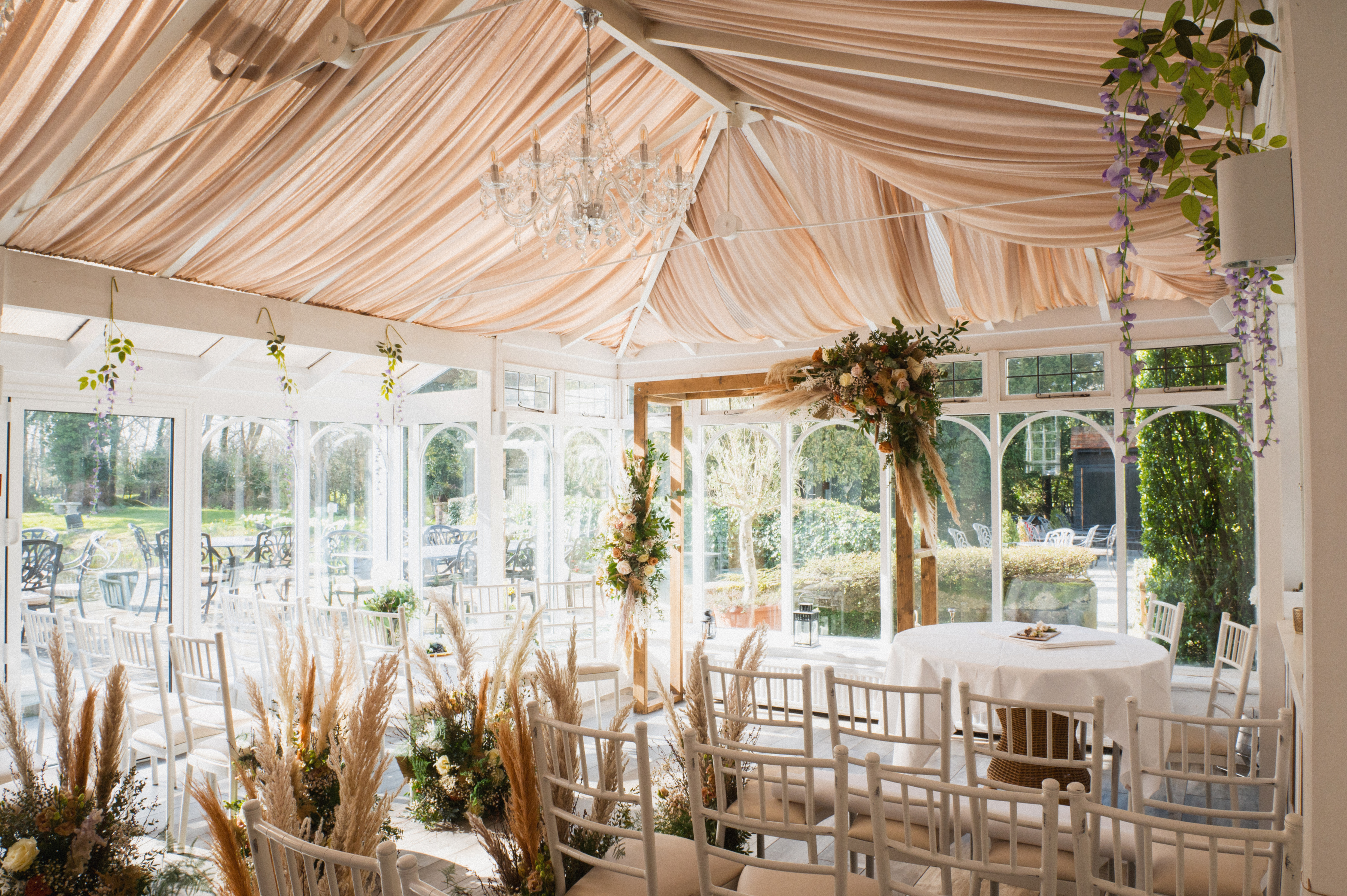 The sunlit conservatory at Sheene Mill set with white chairs, a floral arch and pampas grass, overlooking the gardens, Cambridgeshire