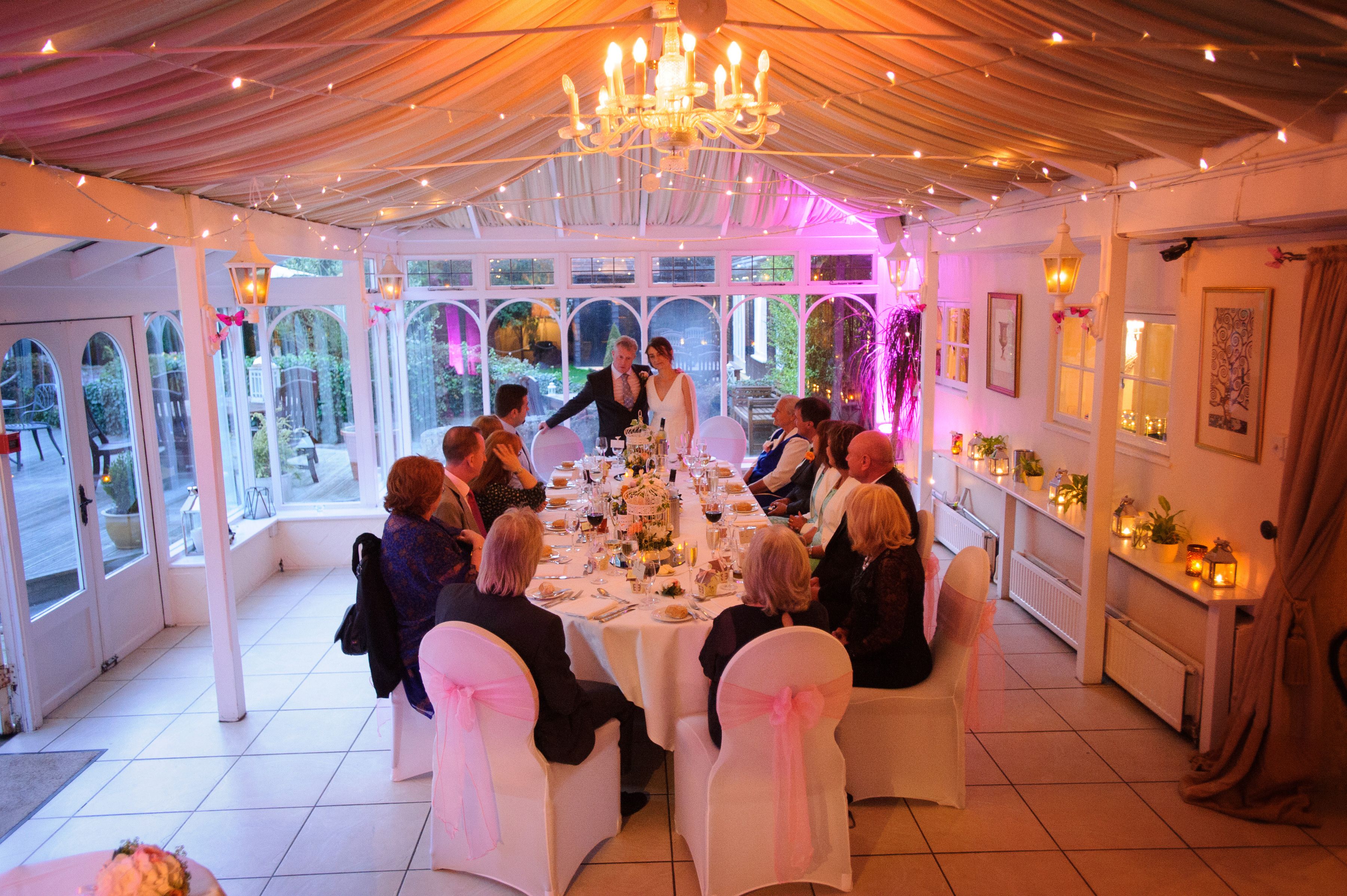An intimate evening wedding dinner in the conservatory at Sheene Mill, a long table under fairy lights and a candle chandelier, Cambridgeshire