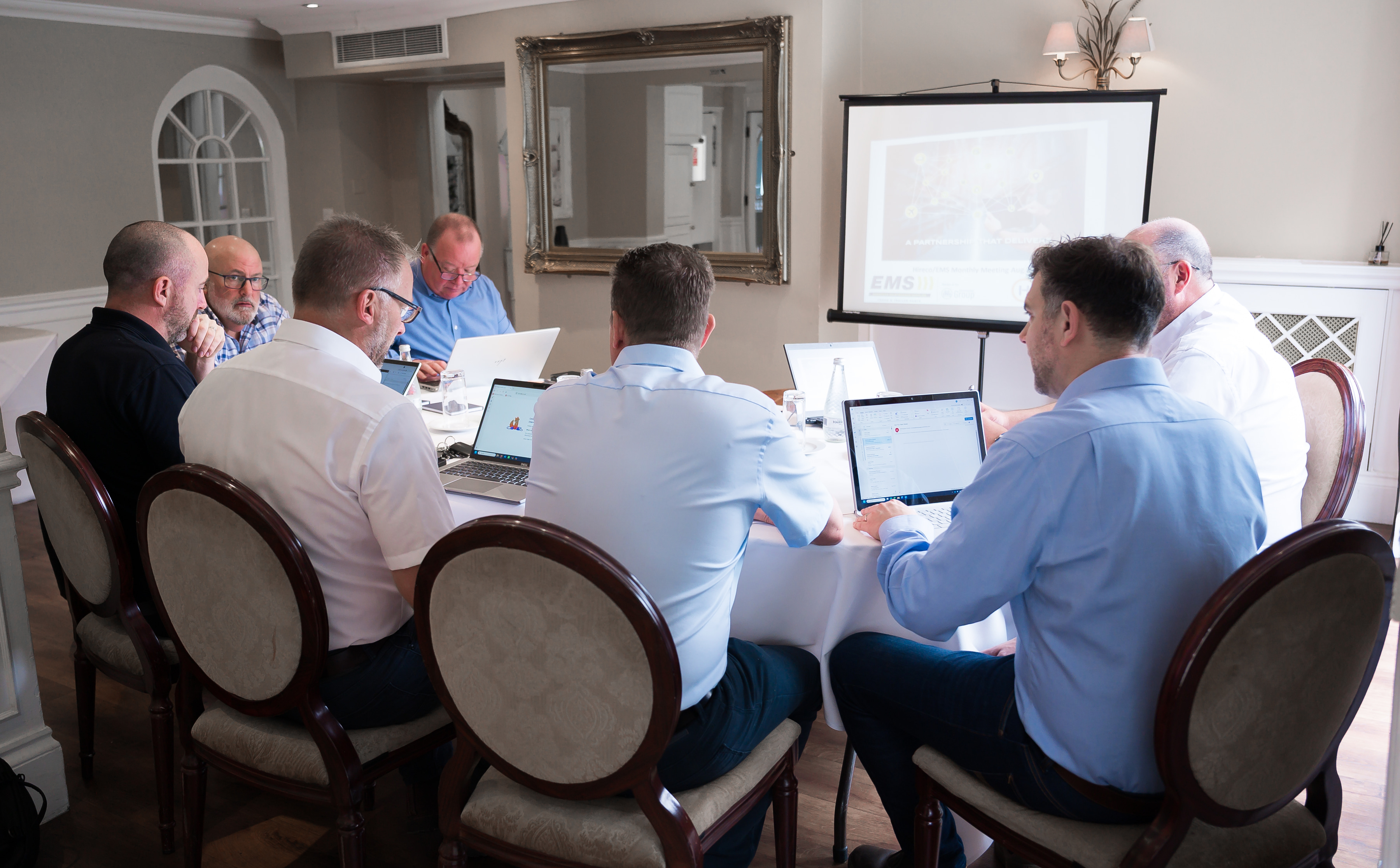 Delegates working on laptops around a round table during a corporate meeting at Sheene Mill, with a presentation on a projector screen beside a gilded mirror, Melbourn, Cambridgeshire