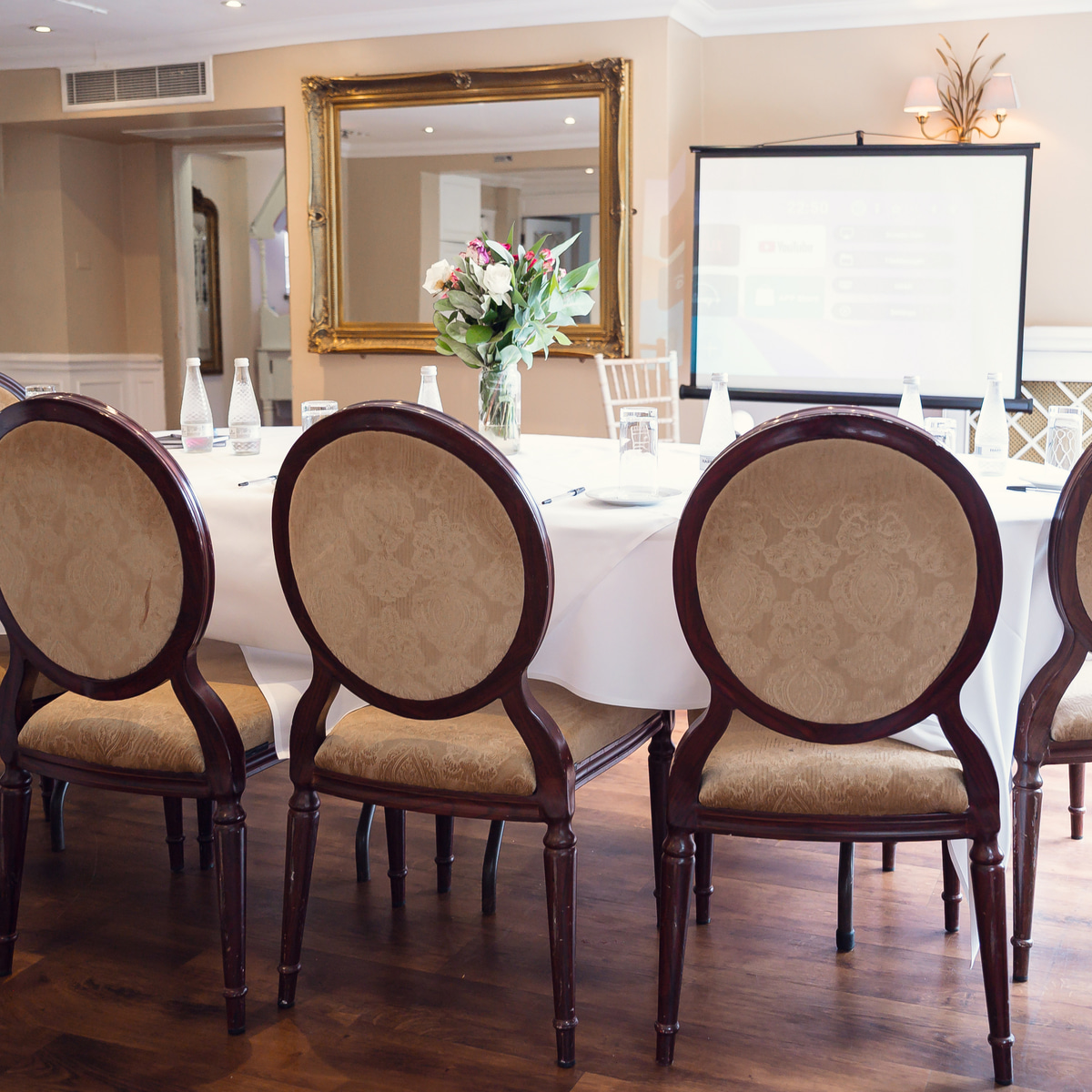 A meeting room at Sheene Mill set for a boardroom meeting, with upholstered chairs around a dressed table, fresh flowers, a gilded mirror and a projector screen