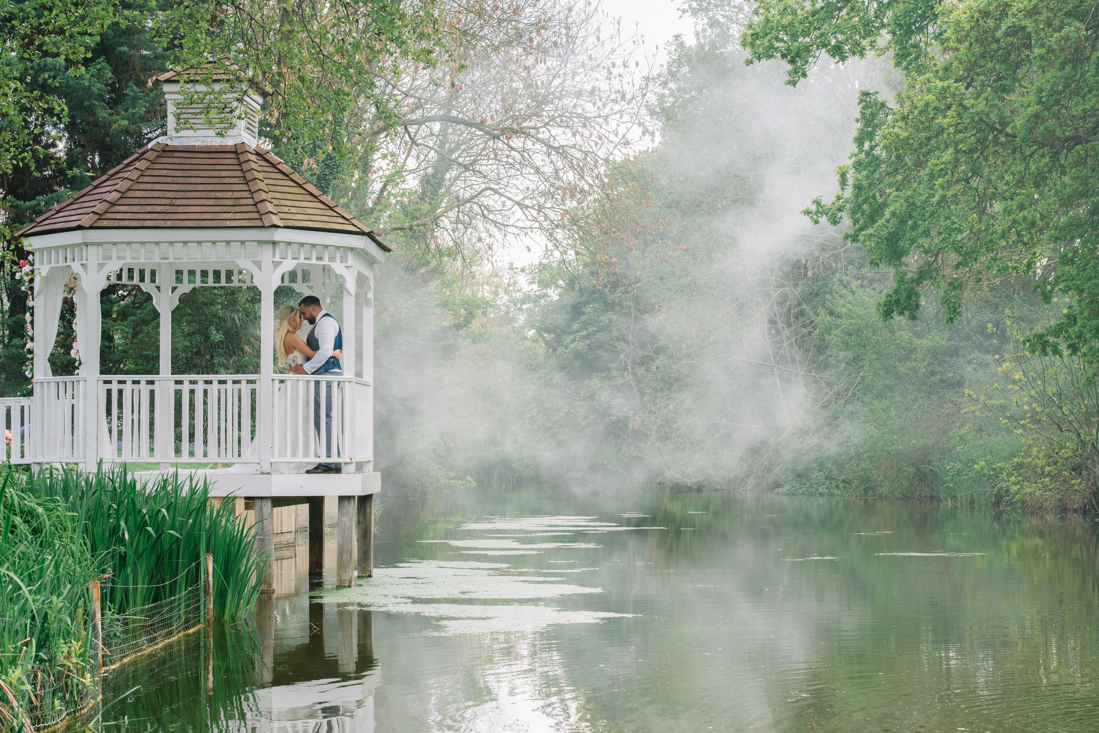 A bride and groom embracing in the white gazebo above the misty mill pond at Sheene Mill, Cambridgeshire