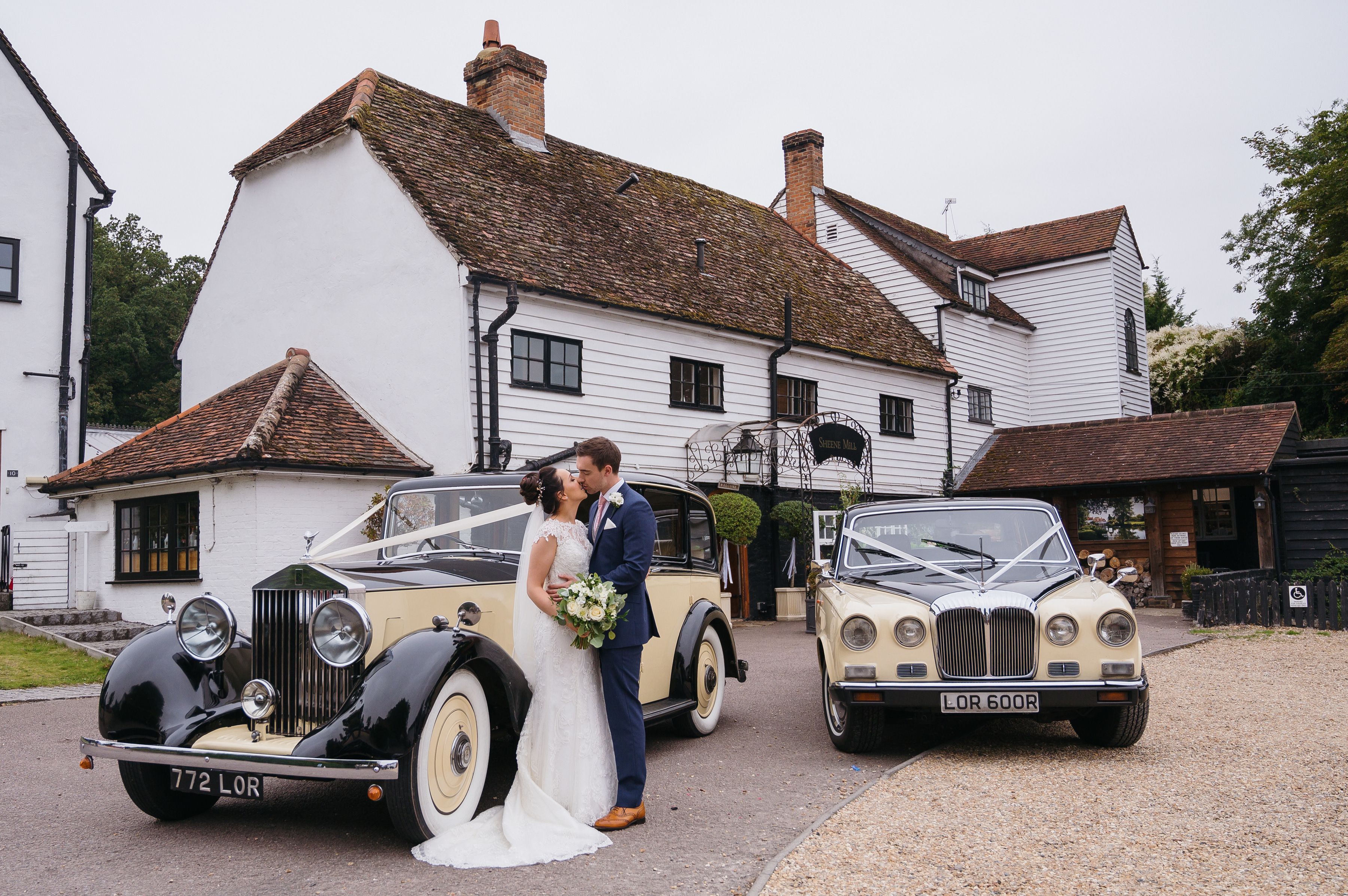 A bride and groom kissing between two vintage wedding cars in front of the mill house at Sheene Mill