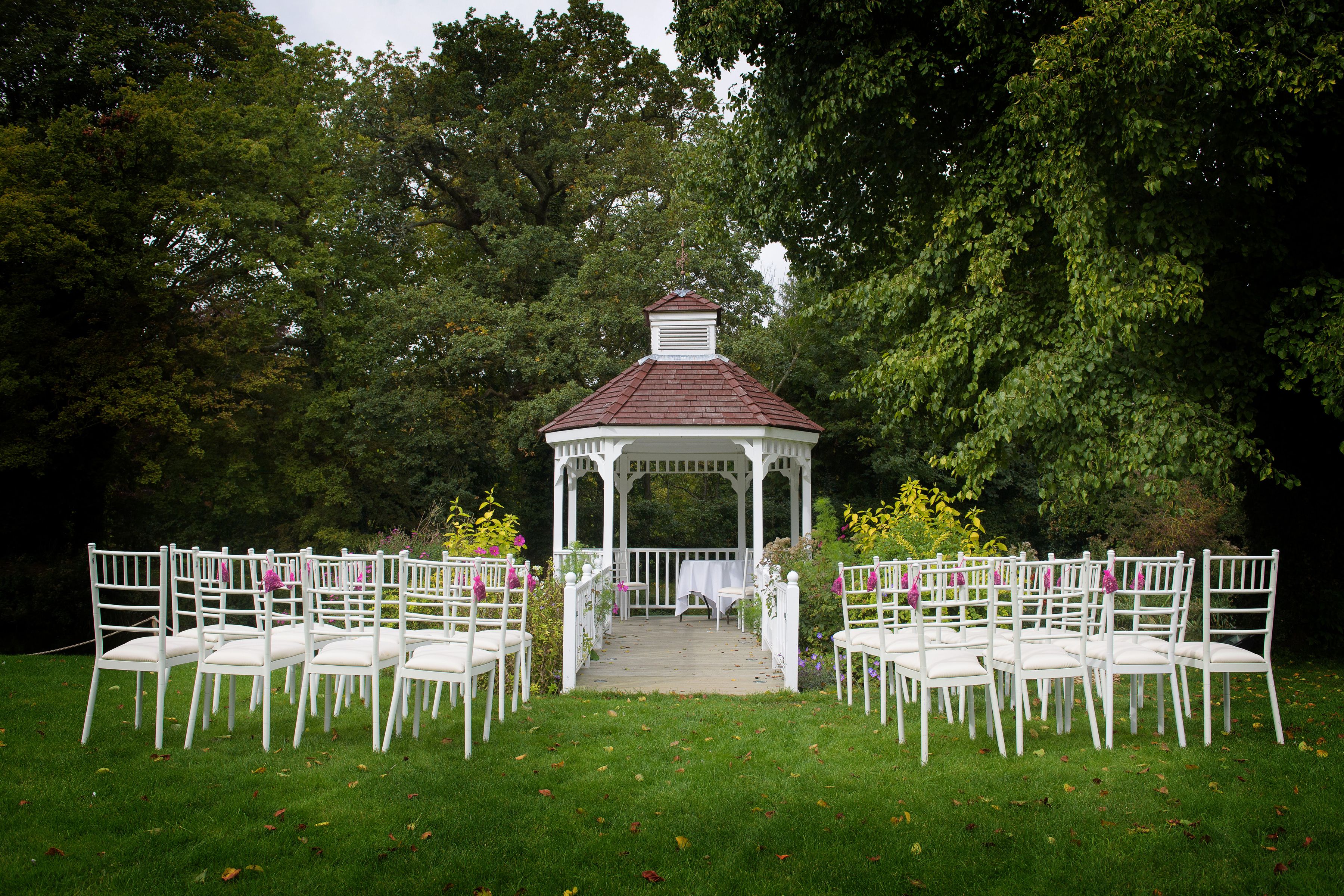 The white garden gazebo set for an outdoor wedding ceremony, with rows of white chairs on the lawn beneath mature trees at Sheene Mill, Cambridgeshire