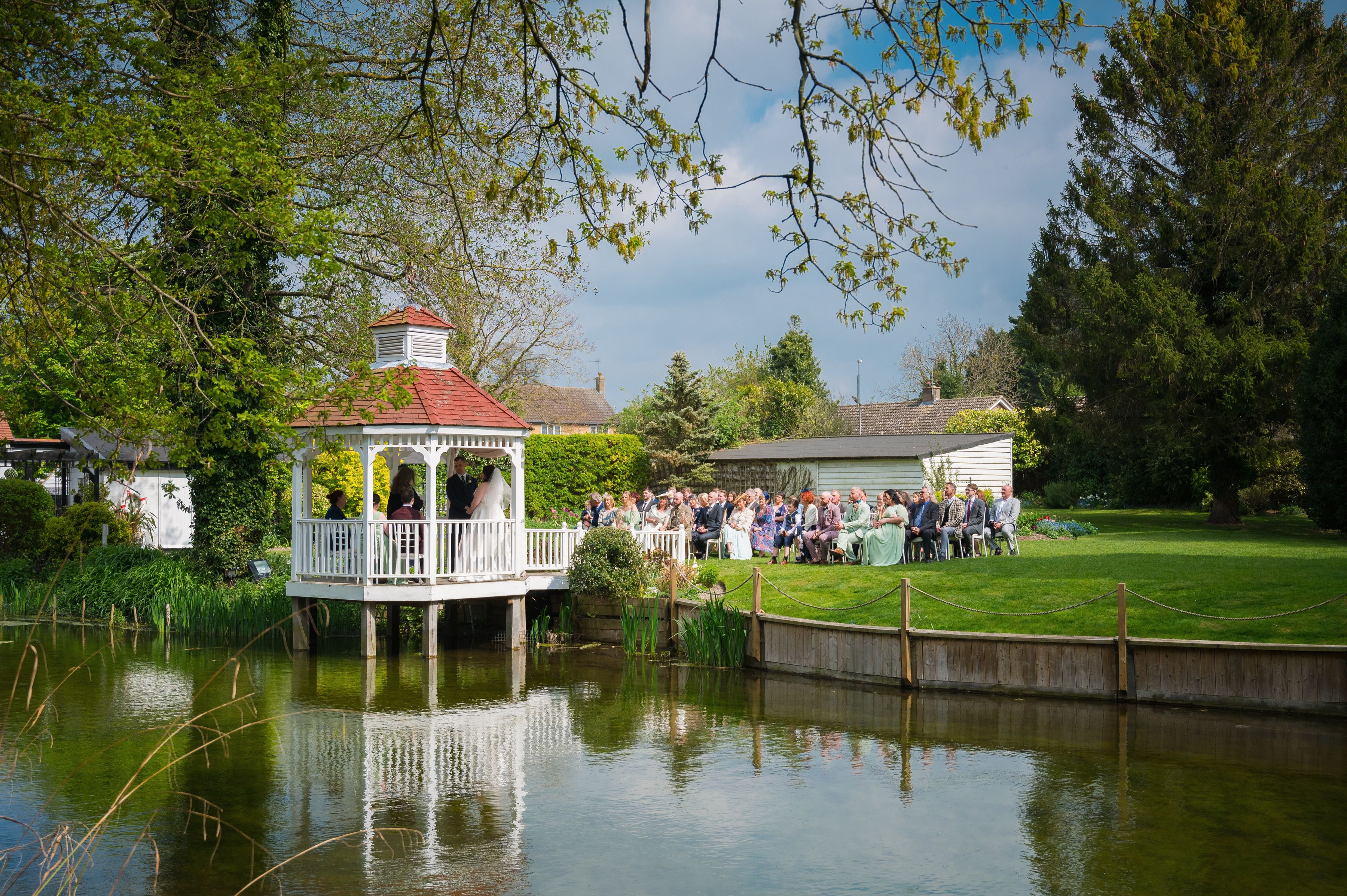 A couple marrying in the white gazebo over the mill pond at Sheene Mill, guests seated on the lawn beside it, Cambridgeshire