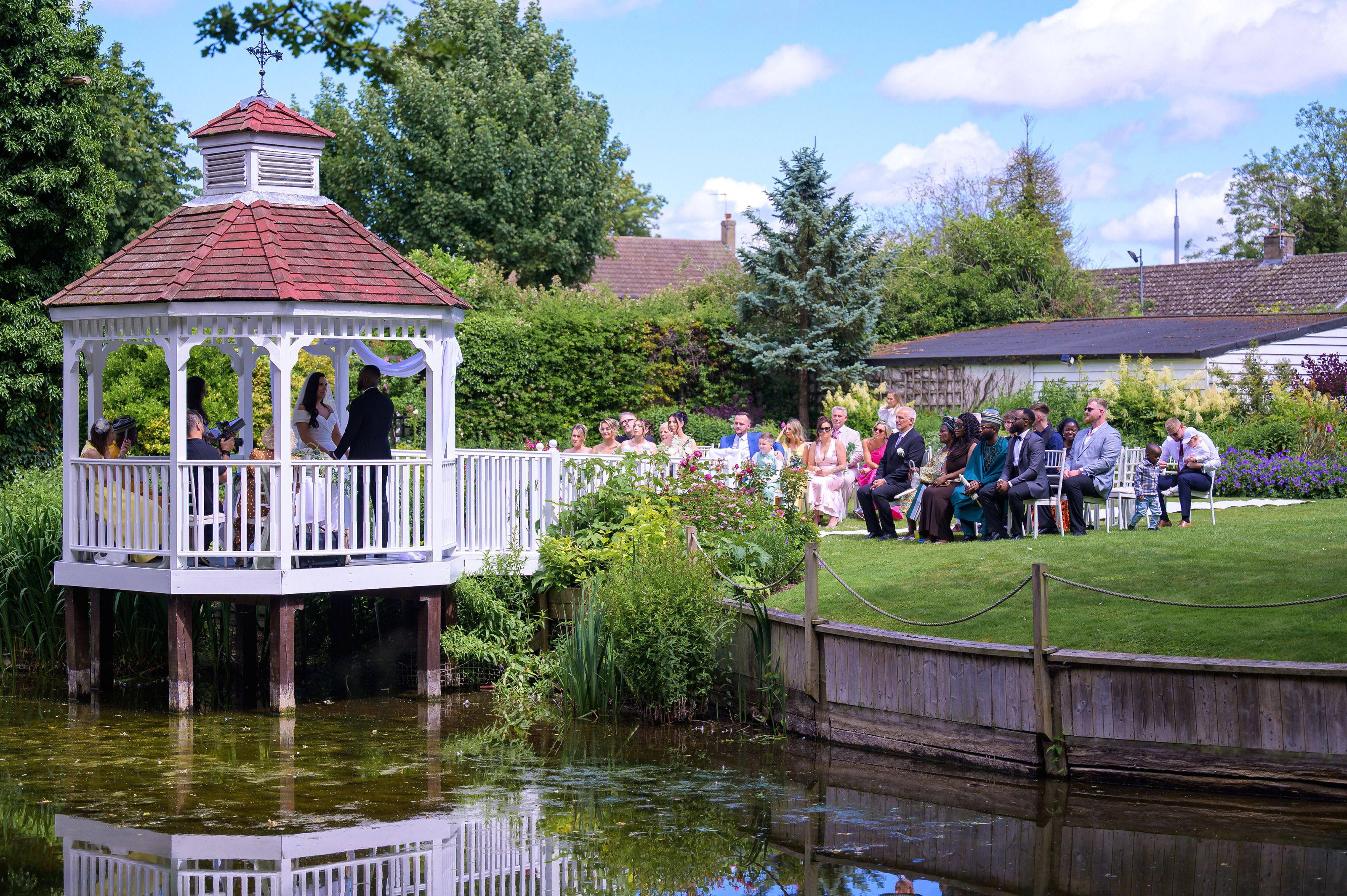 A wedding ceremony beside the gazebo and mill pond at Sheene Mill, guests seated on the riverside lawn, Cambridgeshire
