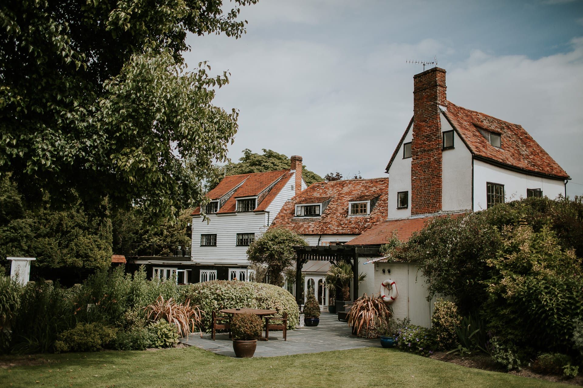 The white weatherboarded mill house at Sheene Mill seen across the garden, with a terrace table set among mature planting, Melbourn near Cambridge
