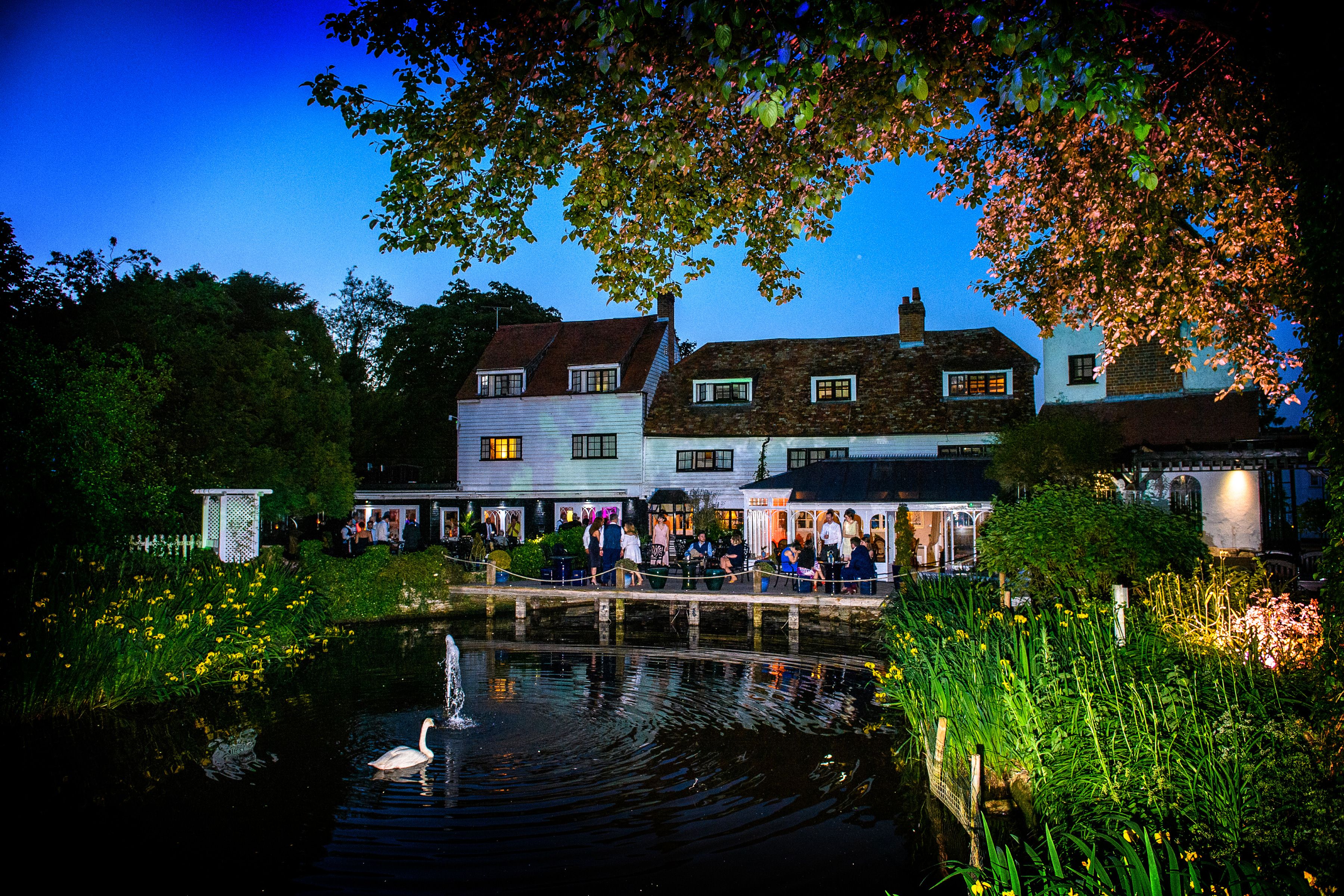 Sheene Mill at dusk across the mill pond, a swan by the fountain and guests gathered on the lit terrace, Melbourn, Cambridgeshire