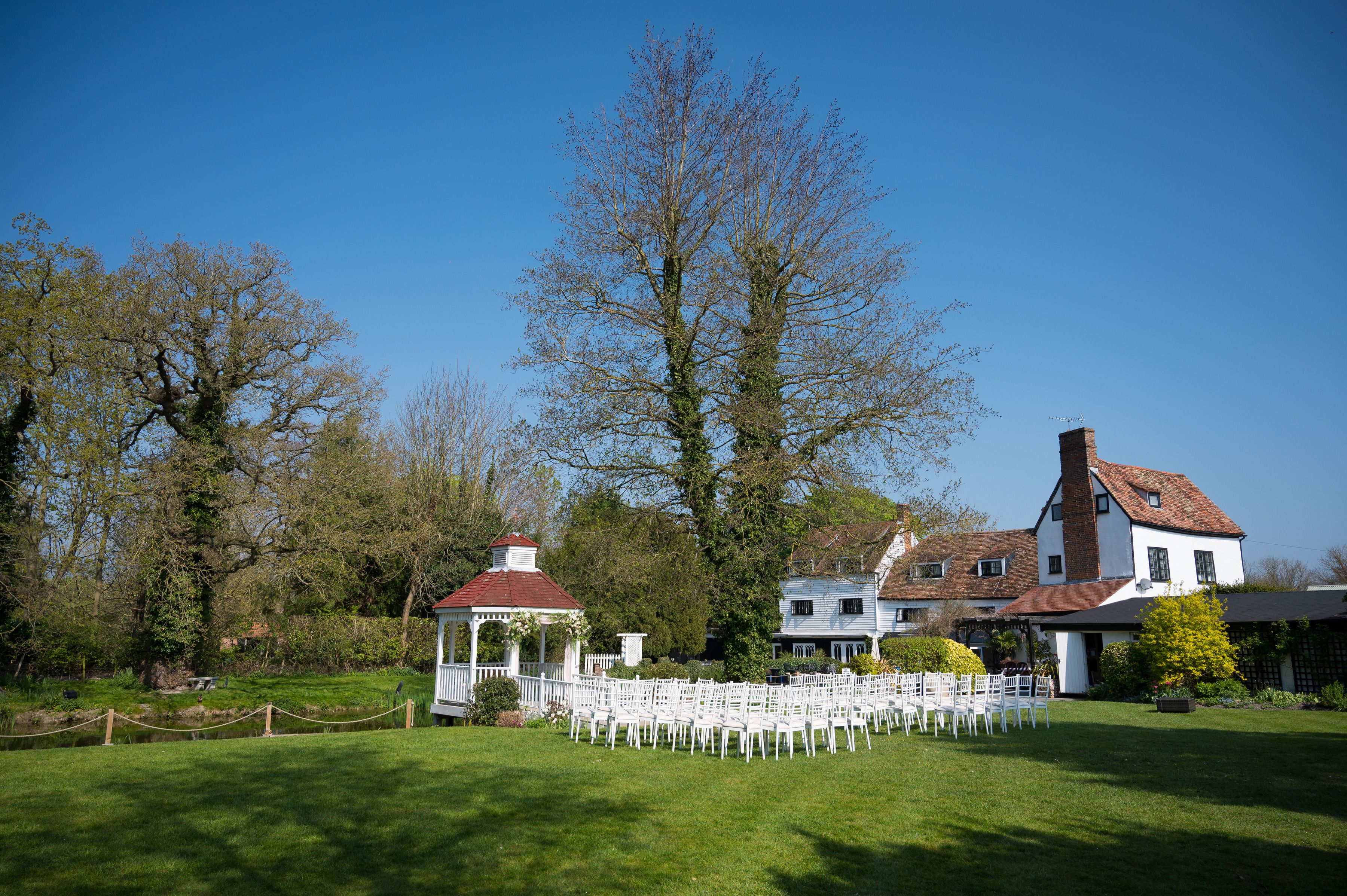 An outdoor ceremony laid out on the lawn beside the gazebo and the white mill house at Sheene Mill, Melbourn, Cambridgeshire