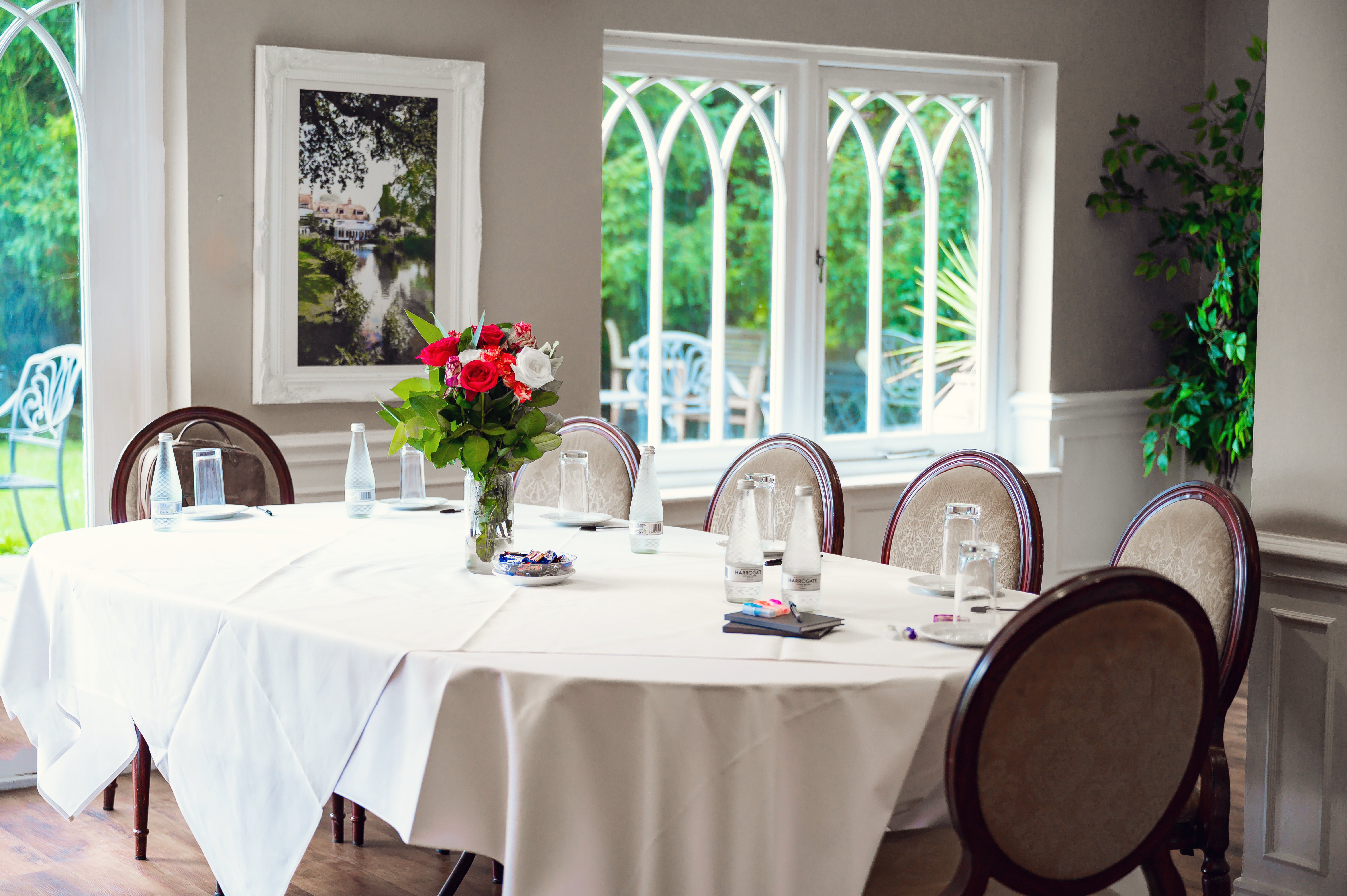 A meeting room at Sheene Mill set for a meeting, with a white-clothed table, red and white roses and arched windows onto the garden, Melbourn, Cambridgeshire