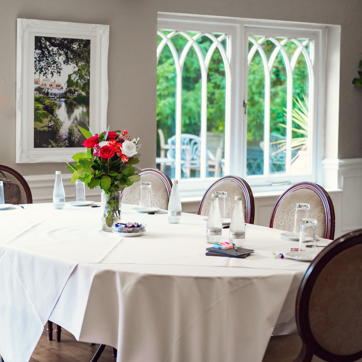 A meeting room at Sheene Mill set for a meeting, with a white-clothed table, red and white roses and arched windows onto the garden