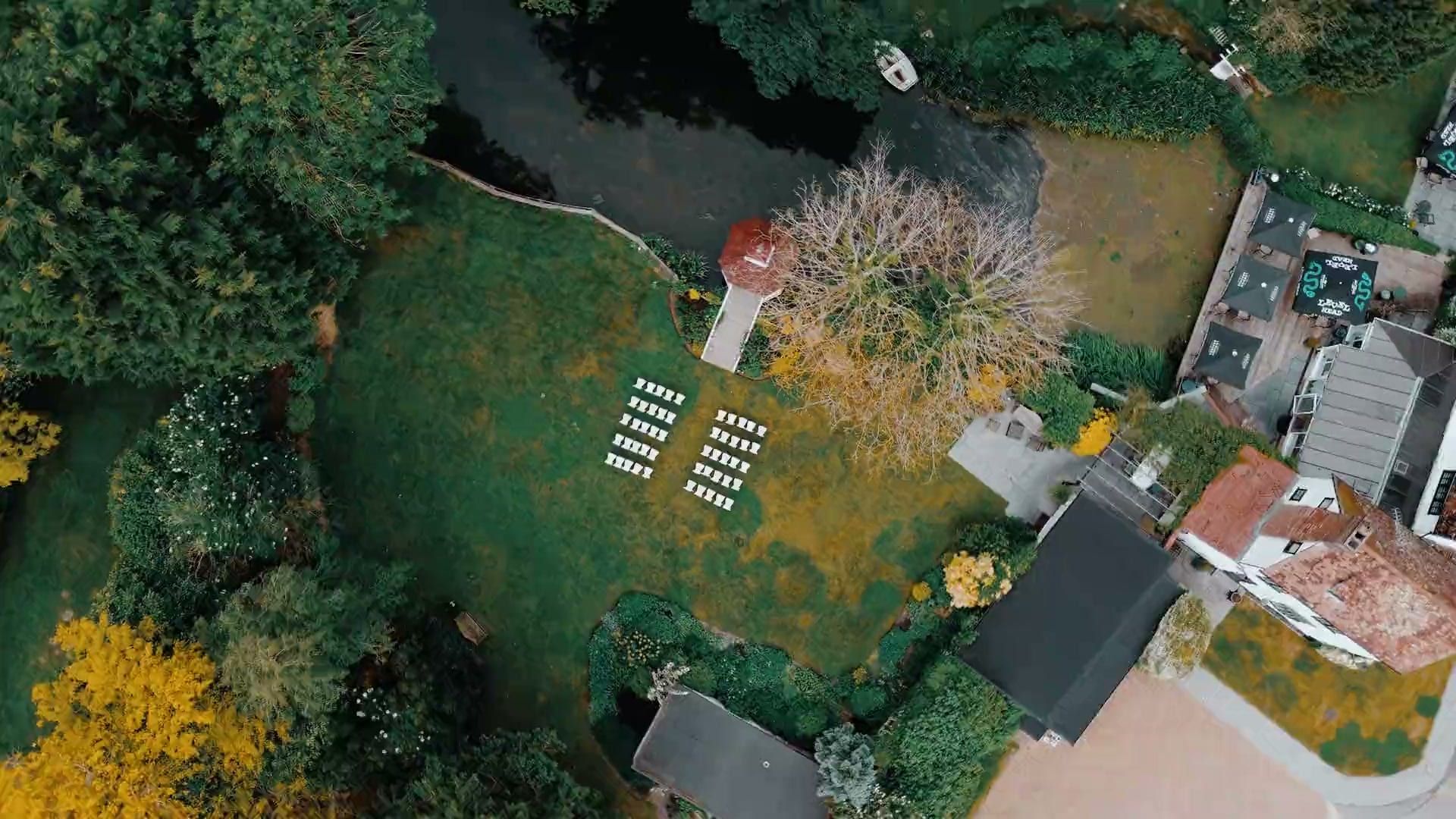 A wedding at Sheene Mill, Cambridgeshire: on phones a ceremony in the white gazebo on the sunlit lawn with guests seated either side, on larger screens an aerial view over the millpond, gazebo and mill house