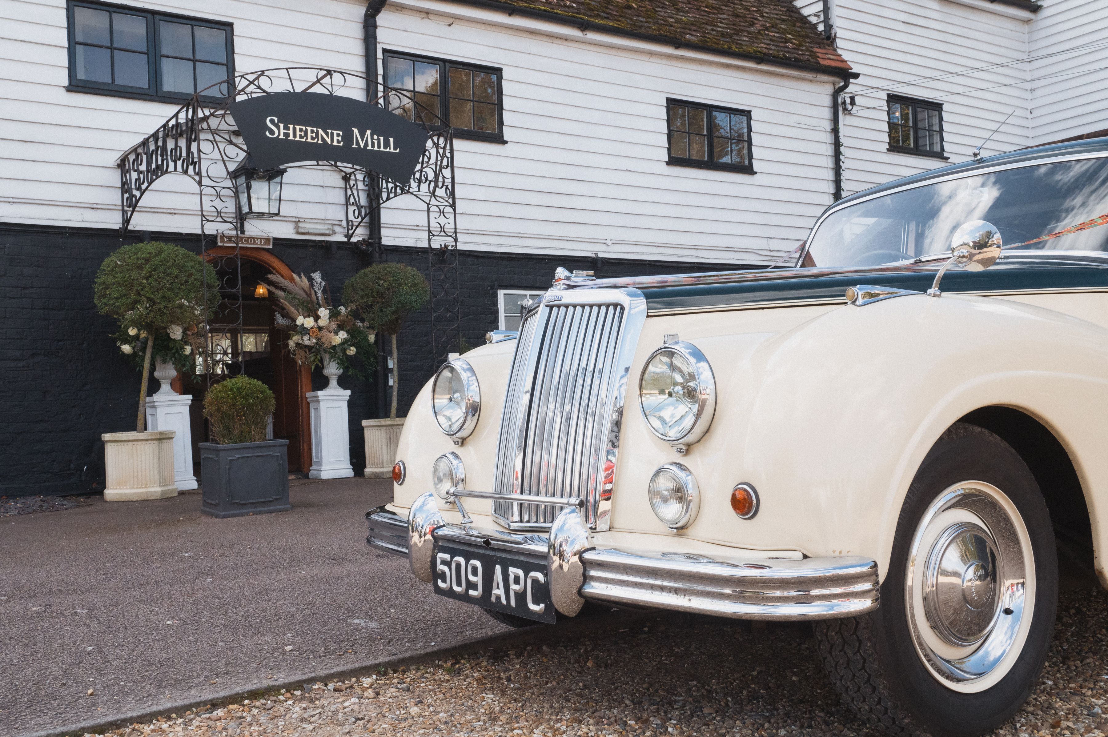A cream vintage wedding car parked at the Sheene Mill entrance beneath the ironwork arch and sign