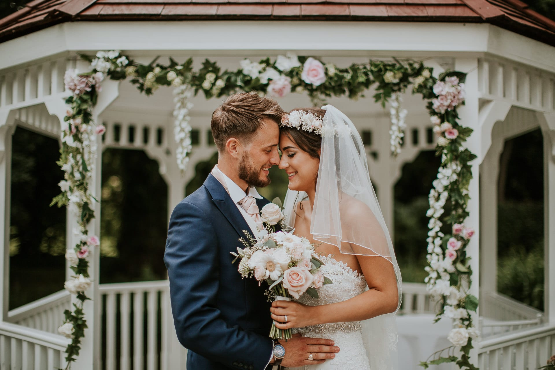 A couple embracing beneath a floral arch on the gazebo at Sheene Mill, the bride holding a pastel bouquet