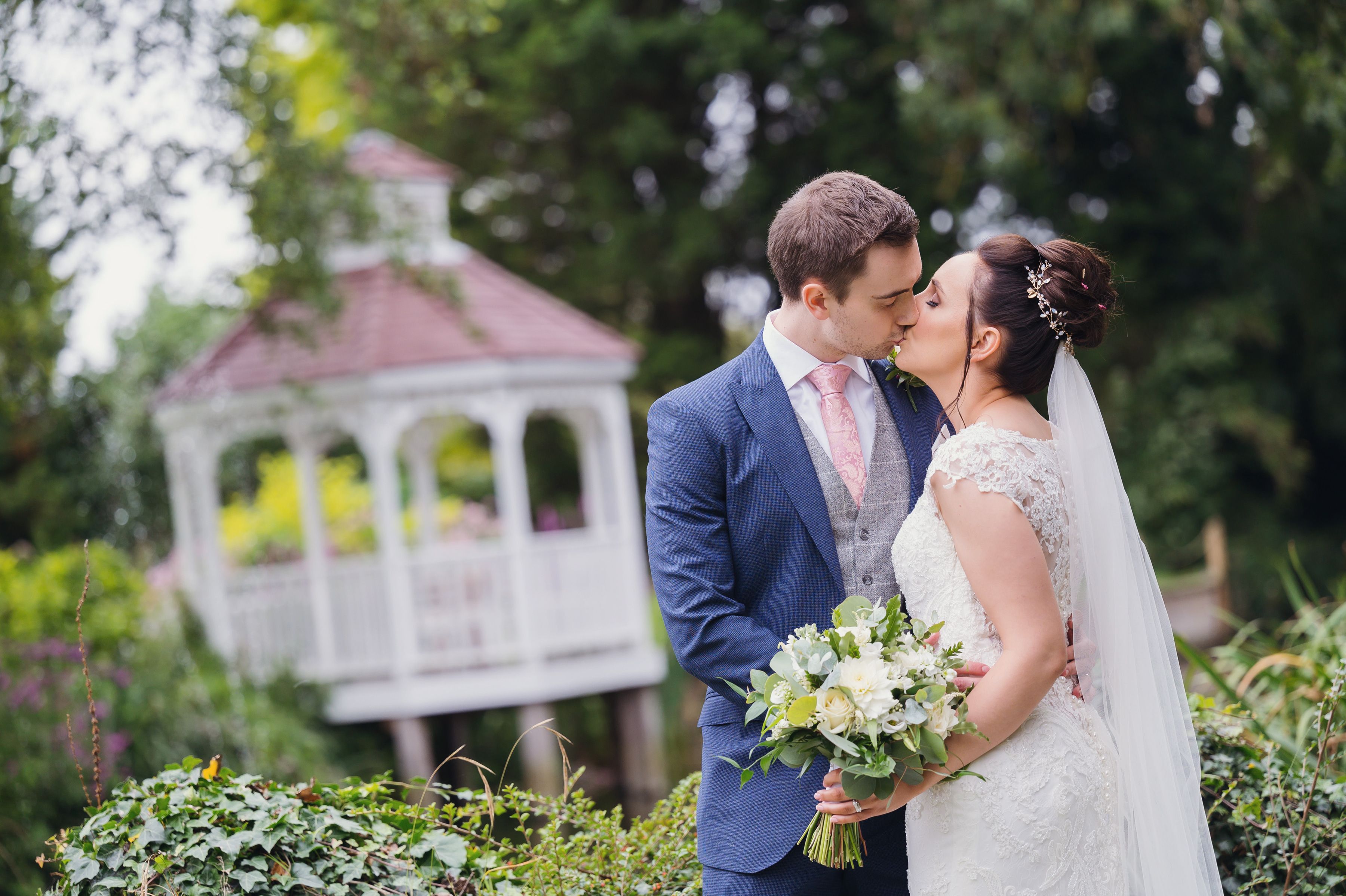 A newlywed couple kissing beside the white gazebo, the bride holding her bouquet, at Sheene Mill