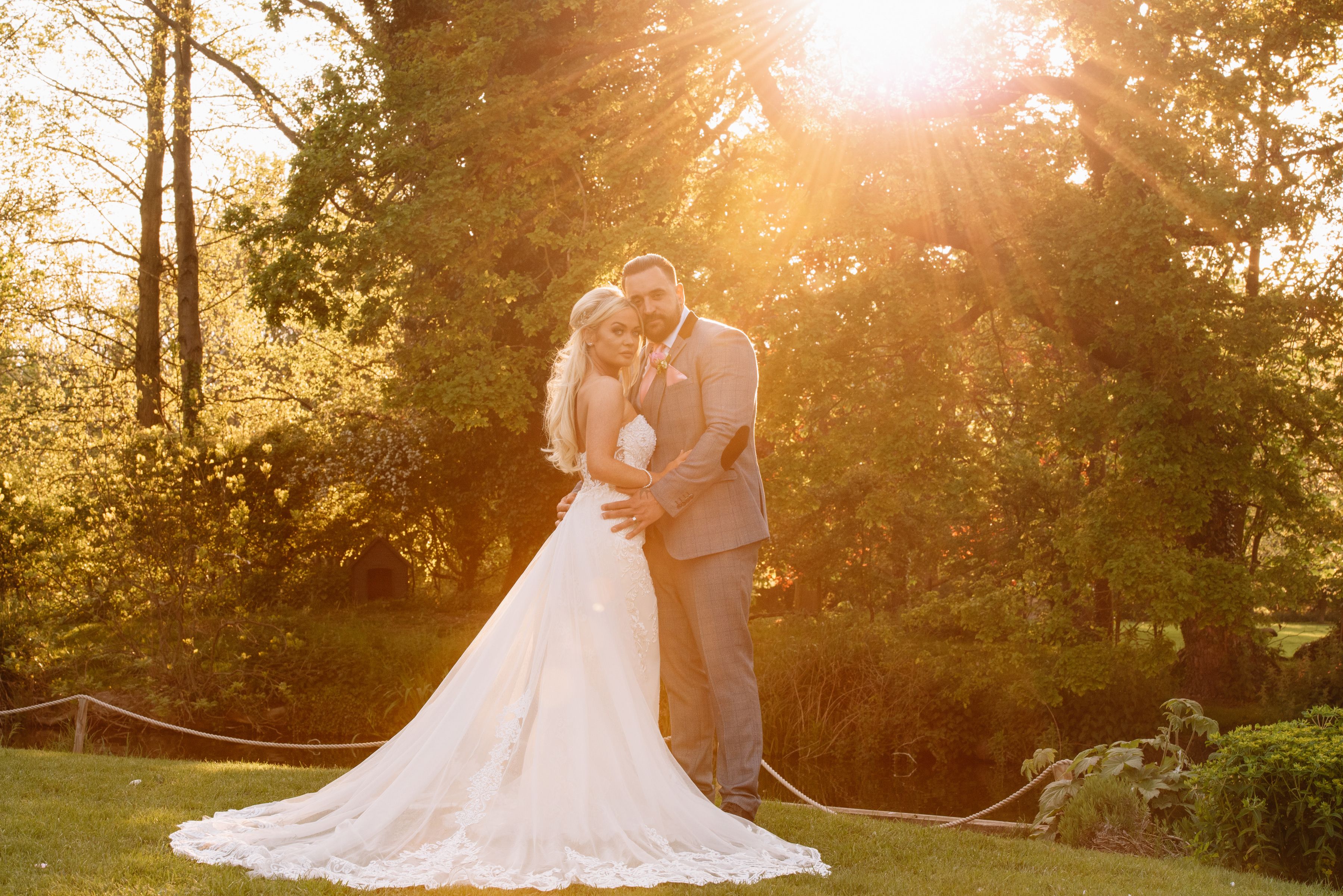 A couple embracing at sunset with light flaring through the trees in the gardens at Sheene Mill